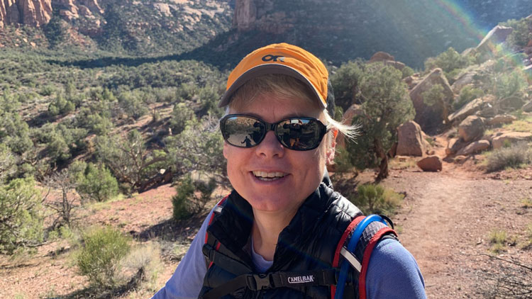 Katharina sits in a high desert landscape, surrounded by pinyon juniper forest and red rock. She wears an orange hat and sunglasses.