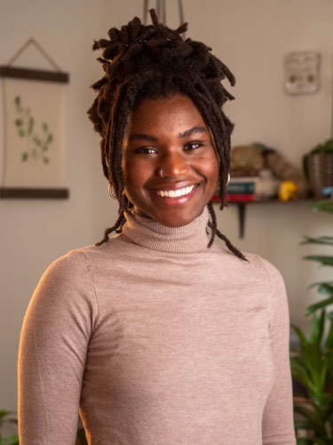 Headshot of Sydney. Her hair is in braids and pulled back in a bun. She's wearing a pink turtleneck. 